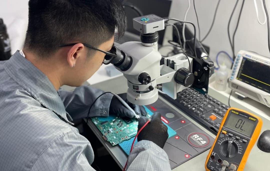 Technician performing component-level microsoldering repair on a circuit board under a digital microscope